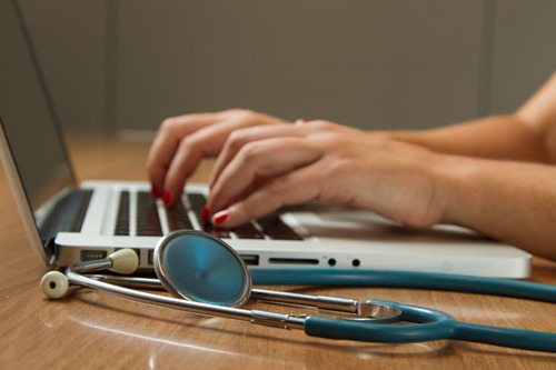 Woman typing on a laptop with a stethoscope on the table.